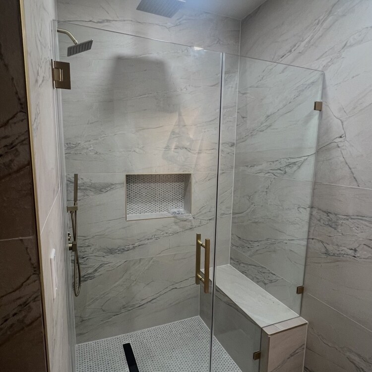 Frameless glass shower with brushed gold hardware beside a marble bench